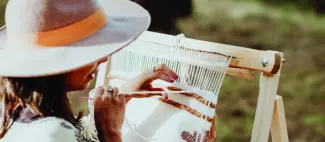 Woman wearing a hat and weaving a mat on a homemade loom in the backyard